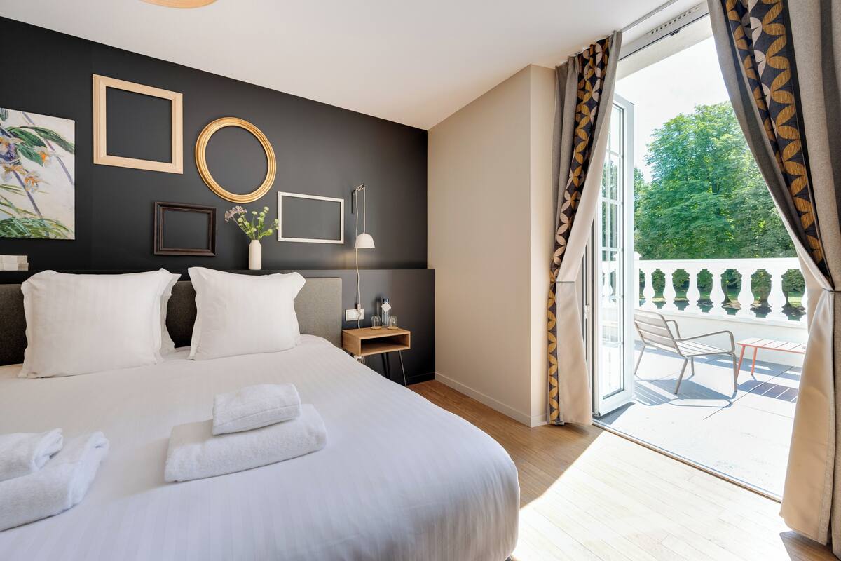 Une chambre d’hôtel avec un grand lit, une table de chevet avec une lampe, une vue sur l’extérieur grâce à une porte coulissante et un balcon avec une chaise.