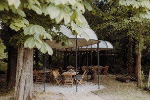 Un espace de restauration en plein air, avec des tables et des chaises disposées sous une grande tente, le tout entouré d’arbres.