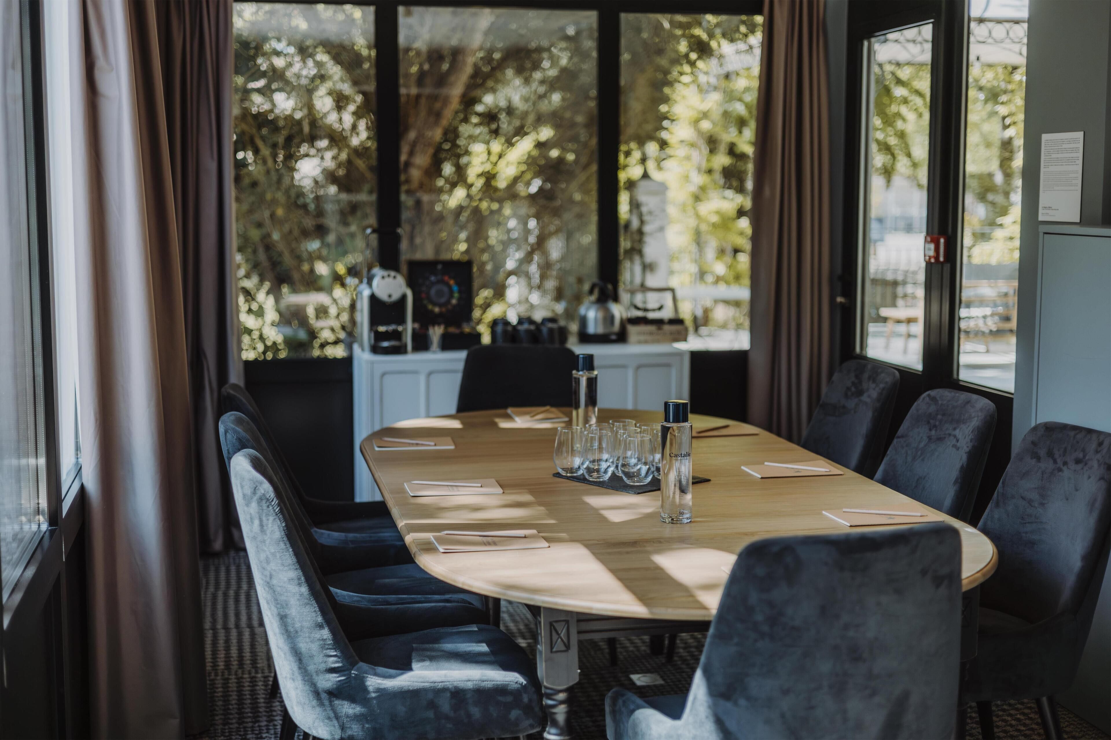 A conference room with a long table, chairs, and a view of trees through large windows.