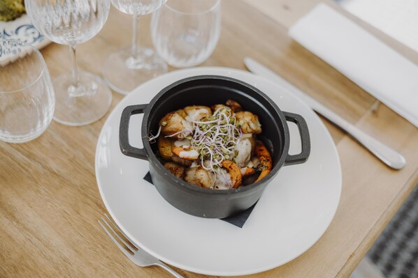 Un plato blanco con una olla negra que contiene comida, un tenedor y copas de vino vacías sobre una mesa de madera.
