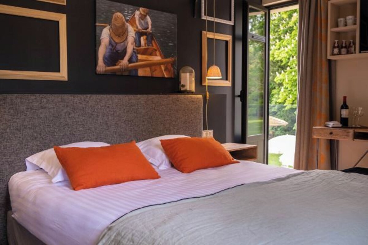 A bedroom with a bed, pillows, a nightstand, a lamp, and a window with a view of greenery.
