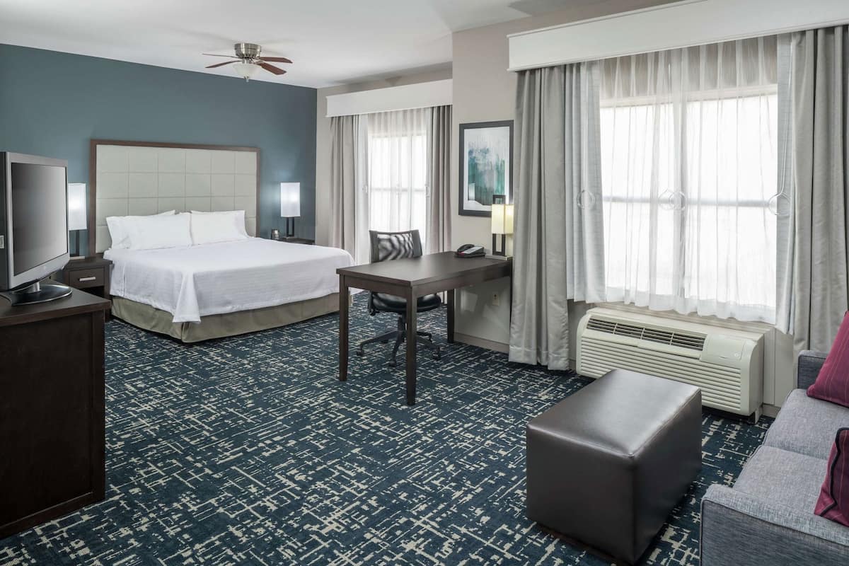 A hotel room with a bed, desk, chair, television, air conditioning unit, and a sofa.