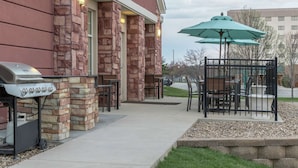 An outdoor patio with stone walls, a grill, and tables with umbrellas. - Homewood Suites by Hilton Cedar Rapids-North (Cedar Rapids)