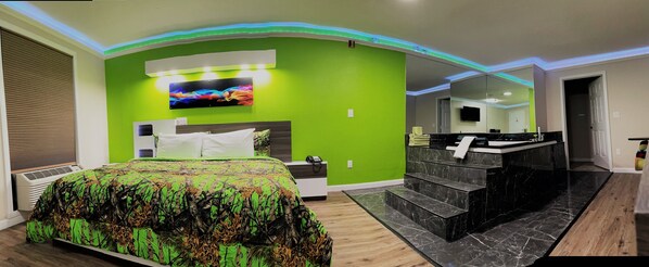 Un dormitorio moderno con una pared verde de acento, una cama grande, un televisor y un baño con encimera de mármol negro.
