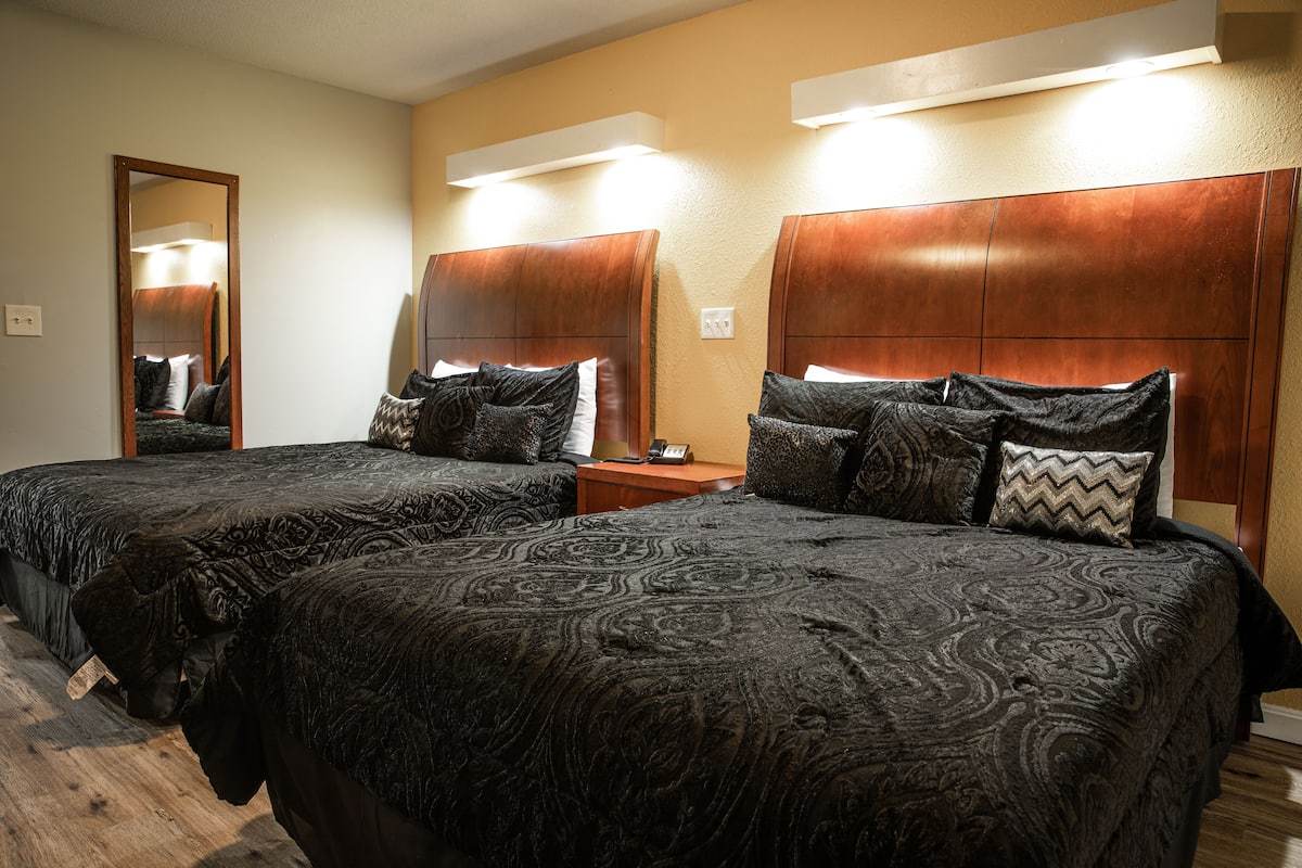 A hotel room with two beds, each with a headboard and multiple pillows. There is a nightstand between the beds with a lamp and a phone.