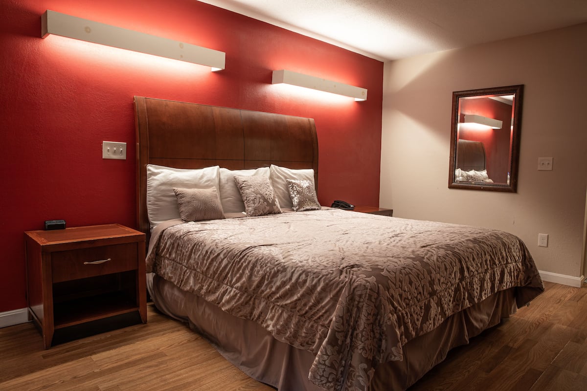 A bedroom with a wooden headboard, a bed with a patterned bedspread, a nightstand, and a wall-mounted mirror.