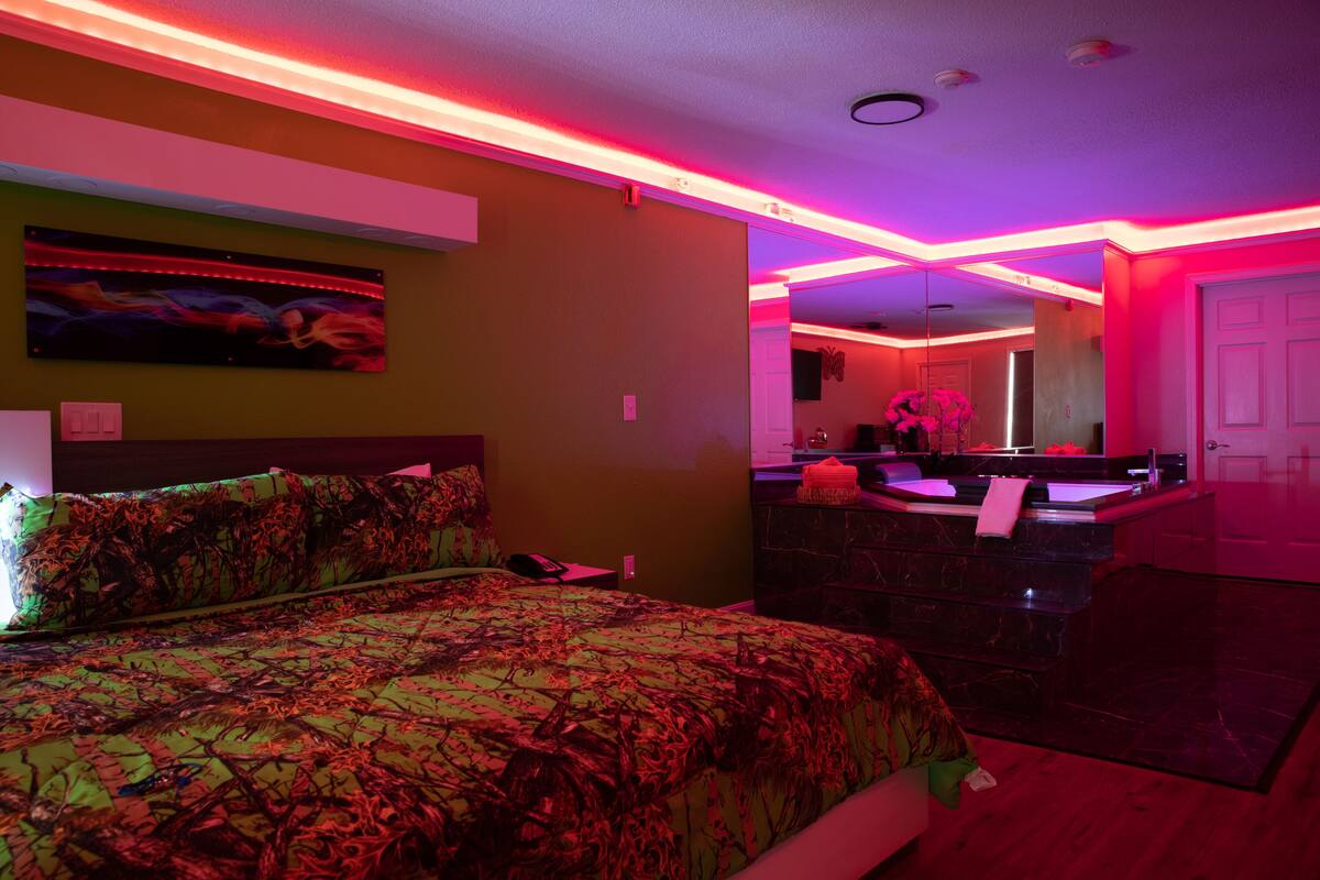 A bedroom with a bed, a nightstand, a mirror, and a bathroom with a sink and mirror.