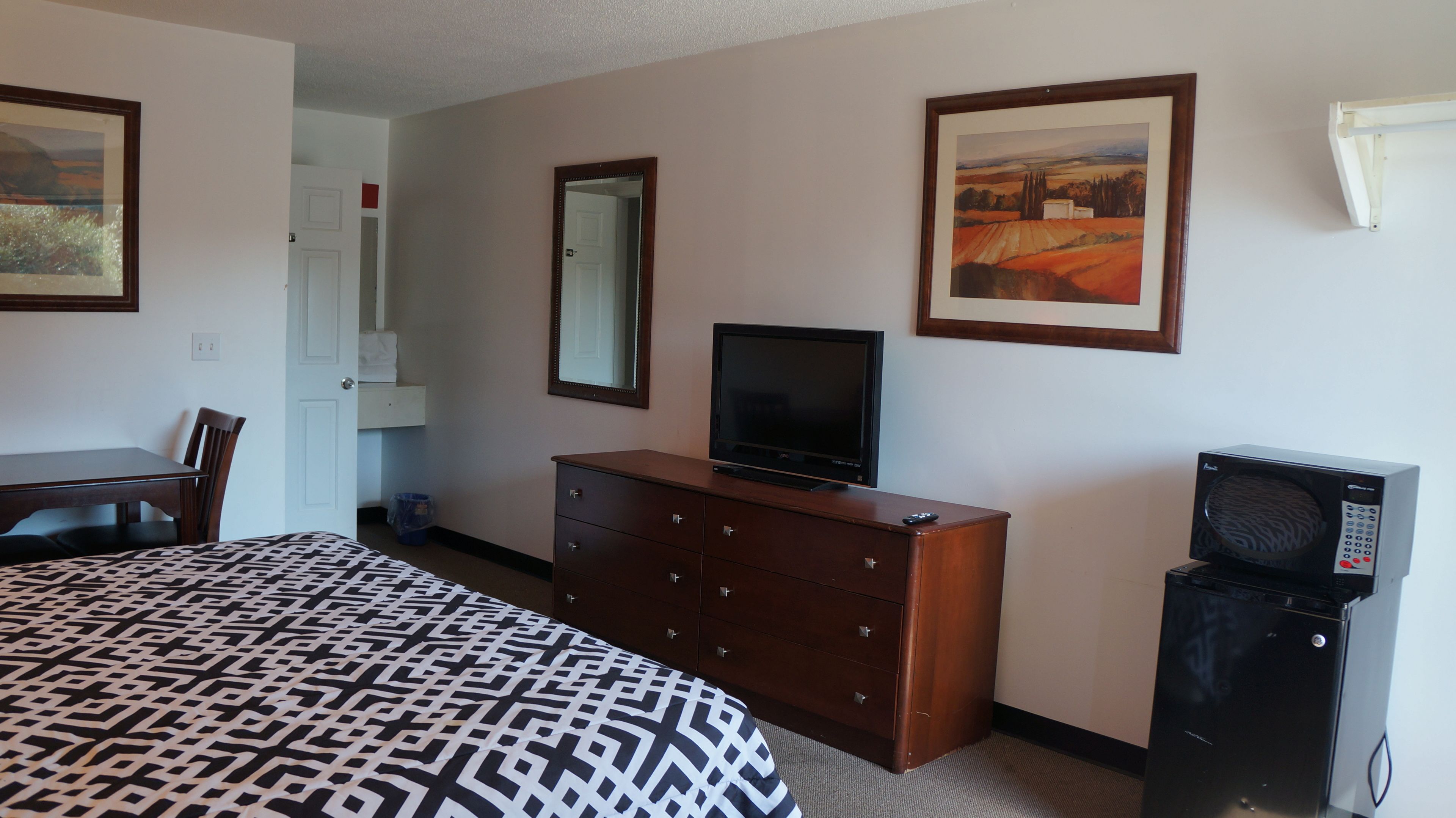 A hotel room with a bed, a dresser, a television, a microwave, a desk, and two framed pictures on the wall.