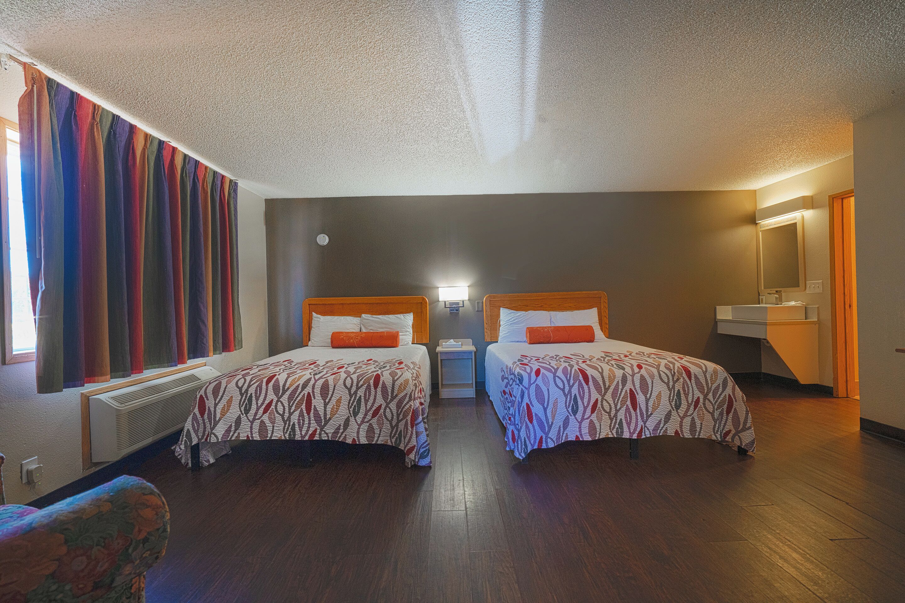 A hotel room with two beds, a large window with colorful curtains, a radiator, and a small bedside table with a lamp.