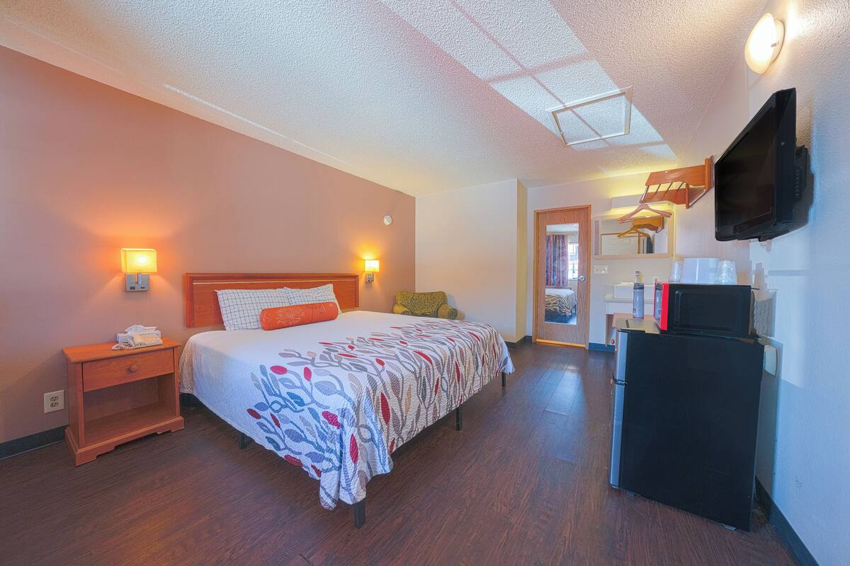 A hotel room with a bed, bedside lamps, a flat-screen TV, a refrigerator, and a wooden nightstand.