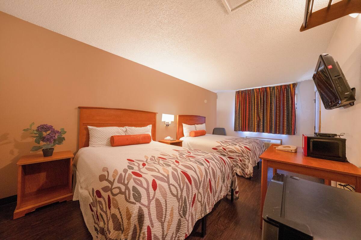 A hotel room with two beds, a wooden nightstand with a lamp, a small table with a microwave, and a television on a wooden stand.