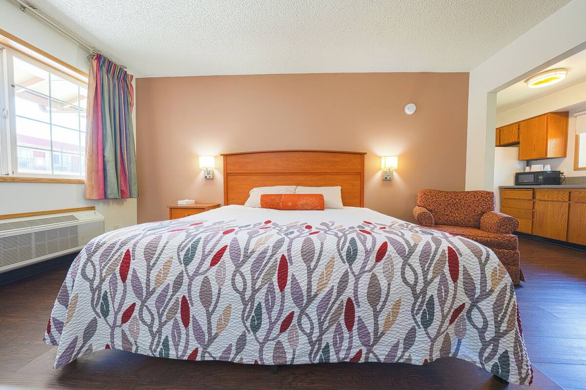 A hotel room with a large bed, a chair, a nightstand, and a view of the kitchen.