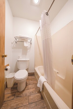 A small bathroom with a toilet, a shower with a curtain, a towel rack, and a trash bin.