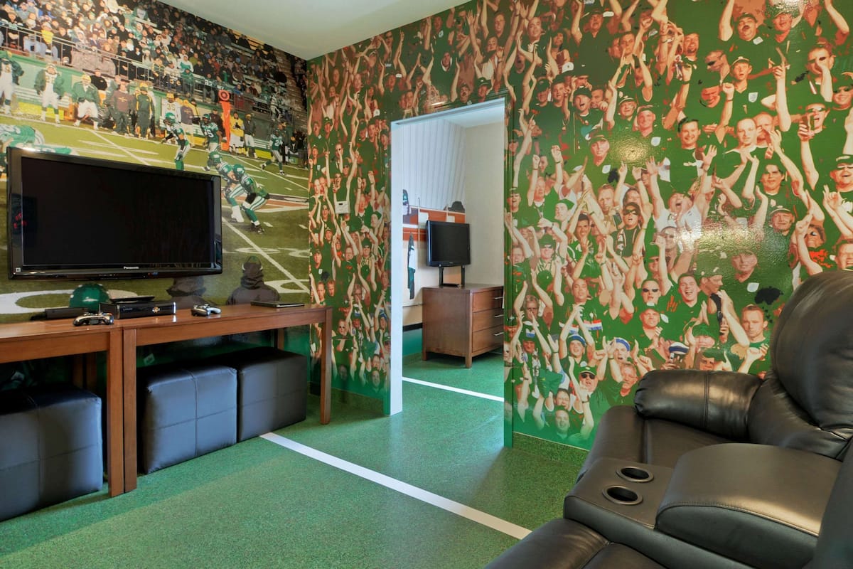 Una habitación con un televisor de pantalla plana grande, un escritorio de madera y un sofá de cuero. Las paredes están cubiertas con un papel tapiz estampado en verde y blanco.