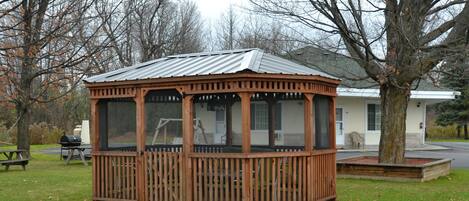A wooden gazebo with a metal roof, surrounded by a grassy area and trees.