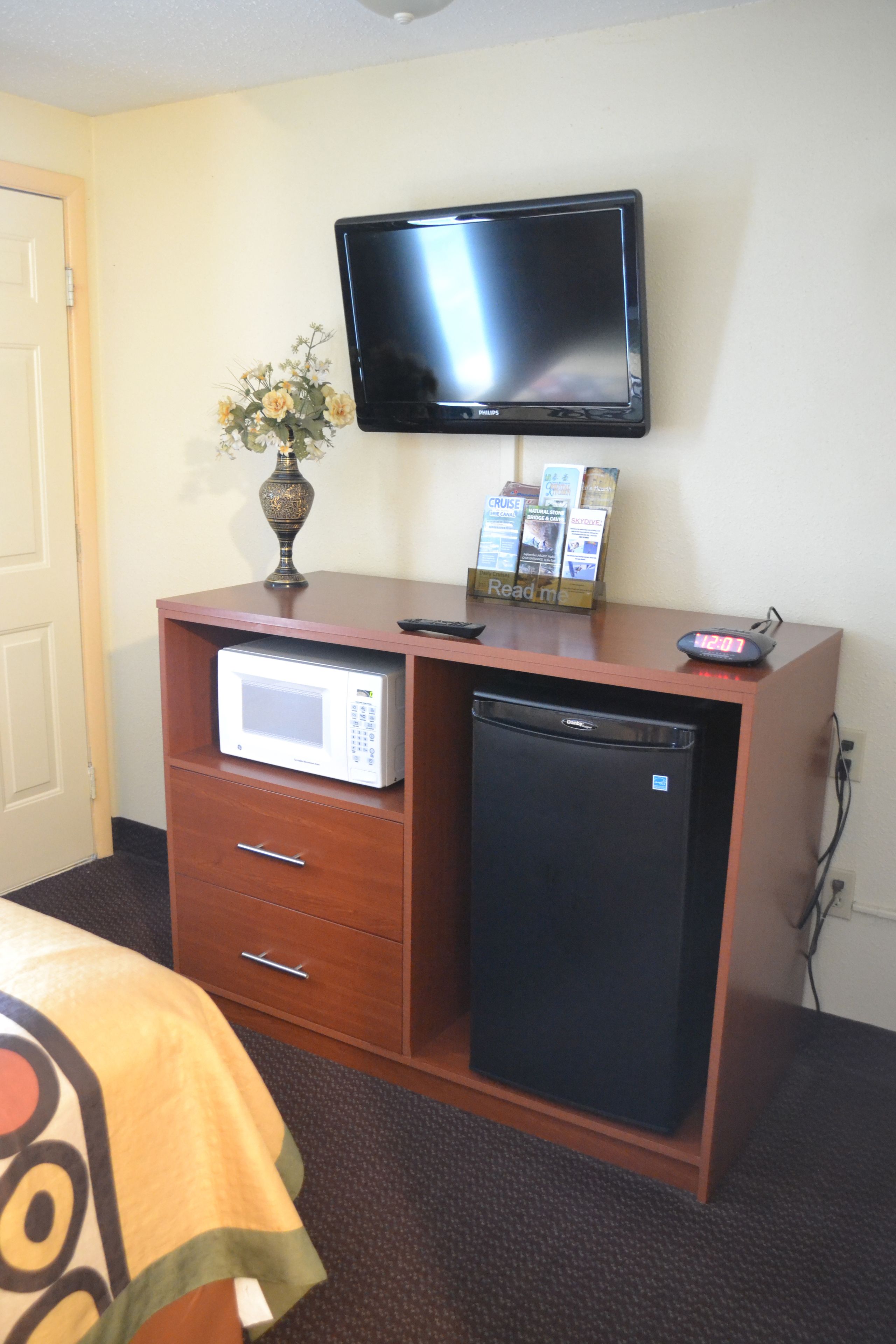 A hotel room with a wooden desk, a microwave, a refrigerator, a television mounted on the wall, and a vase with flowers.