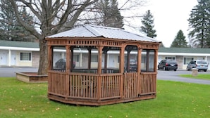 Un gazebo in legno con tetto in metallo, circondato da erba e alberi, di fronte a una fila di edifici in stile motel.