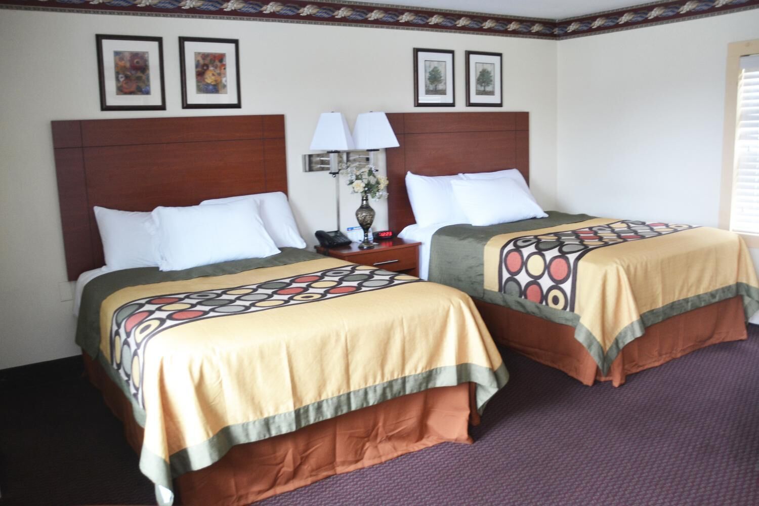 Una habitación de hotel con dos camas, cada una con una colcha estampada, cabecero de madera, mesita de noche con lámpara y cuadros en la pared.