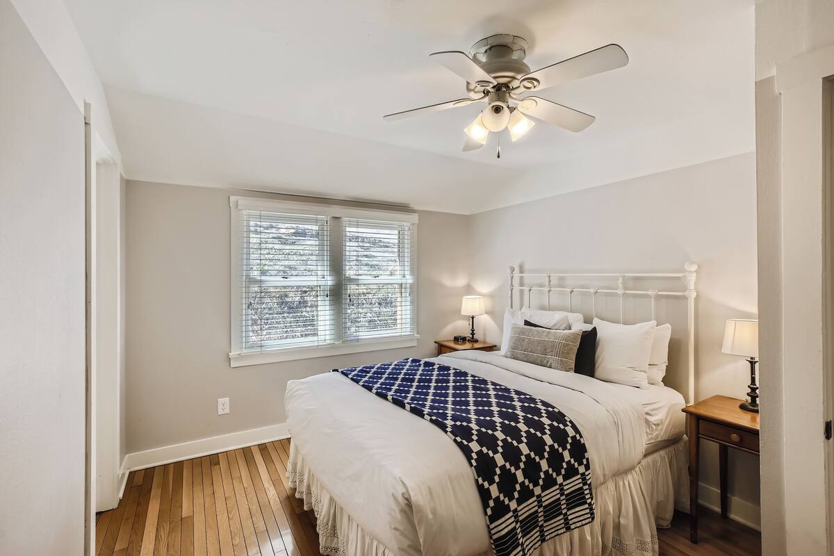 A bedroom with a white bed, a ceiling fan, a window with blinds, and a nightstand with a lamp.