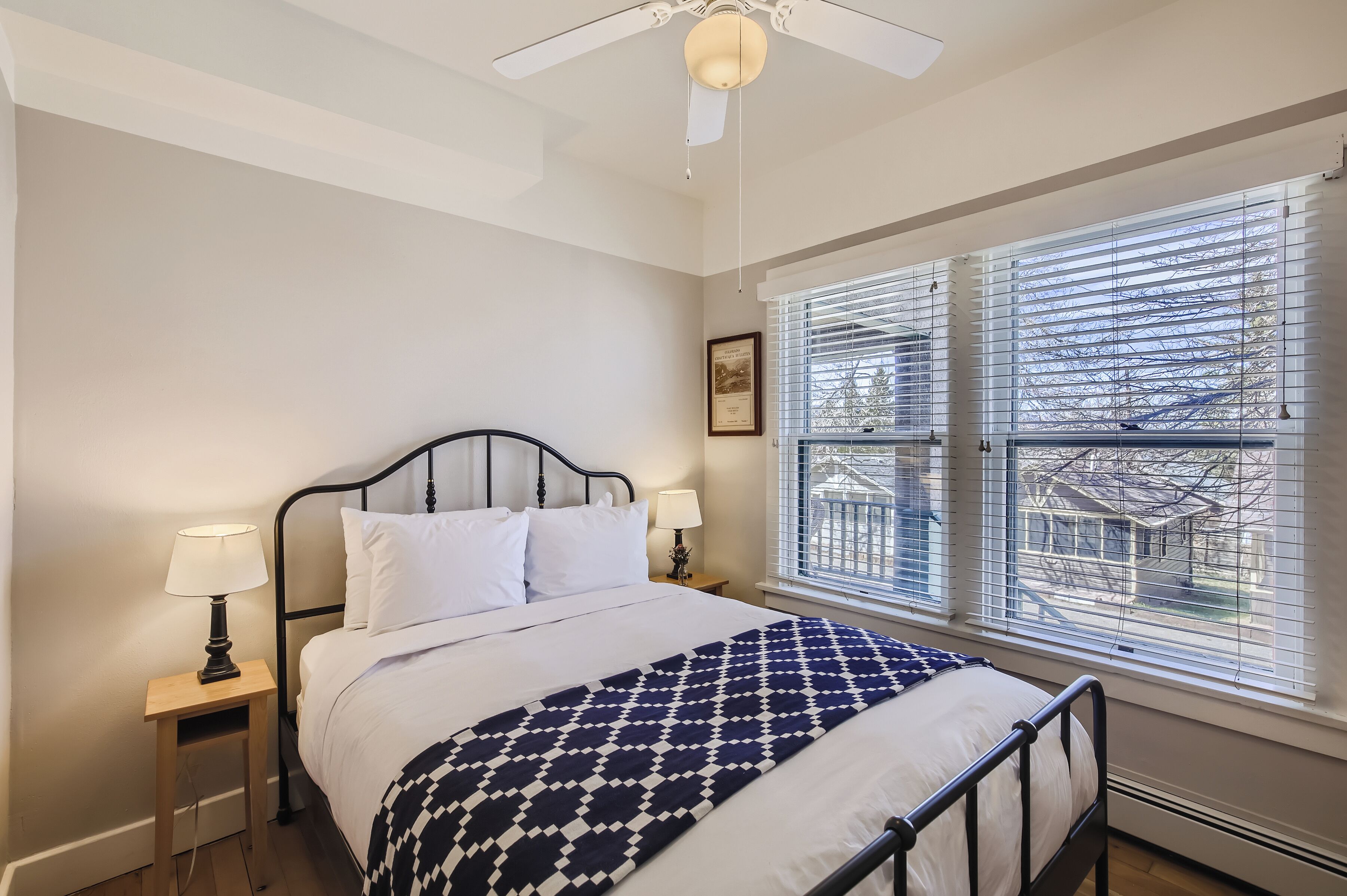 Quarto com uma cama, duas luminárias de cabeceira, um ventilador de teto e uma janela grande com persianas.
