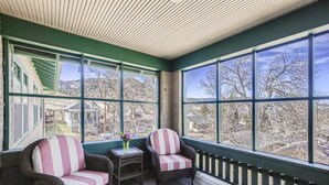 A covered porch with wicker chairs and a small table, large windows offering a view of a residential area. - Colorado Chautauqua (Boulder)