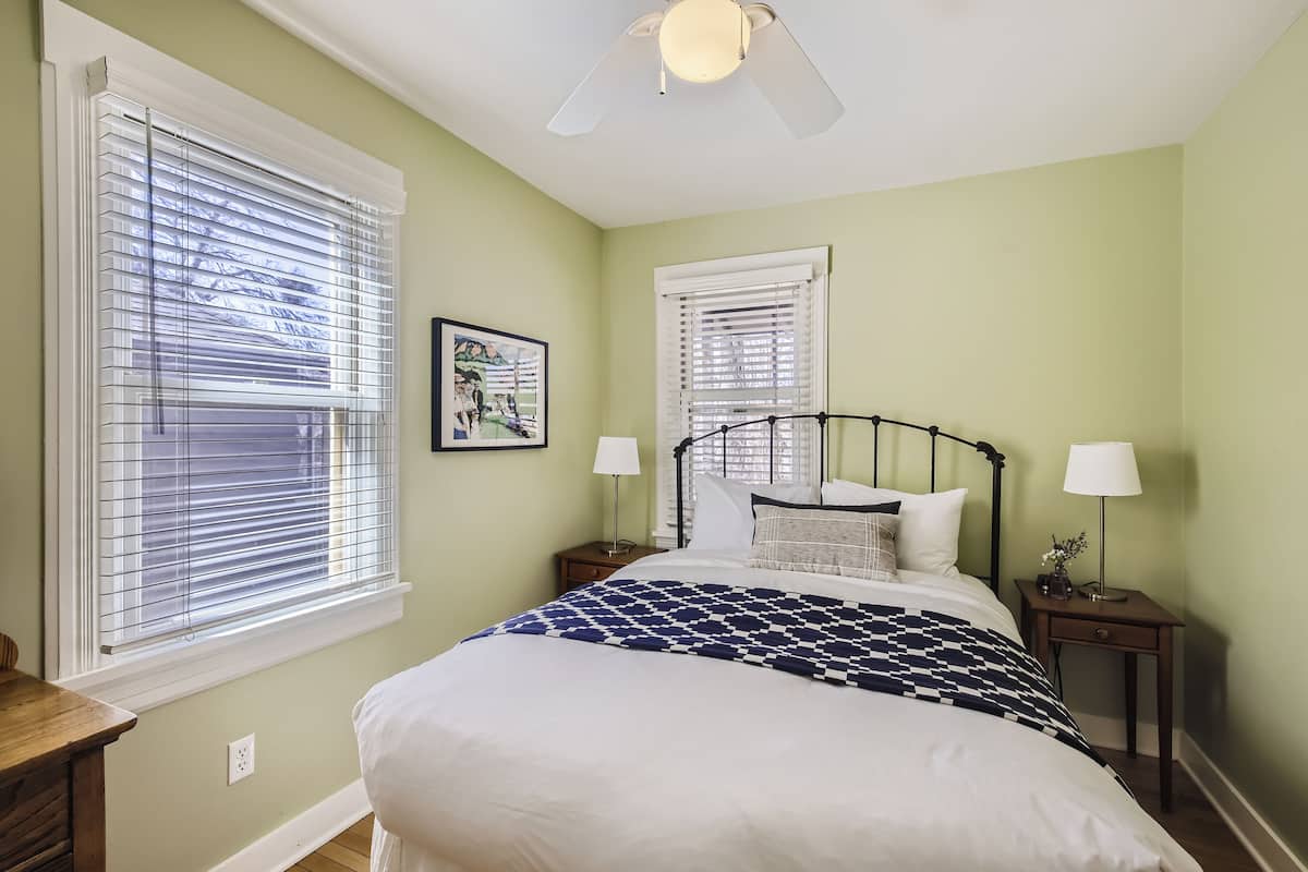 A bedroom with a bed, two nightstands, a ceiling fan, and two windows with blinds.