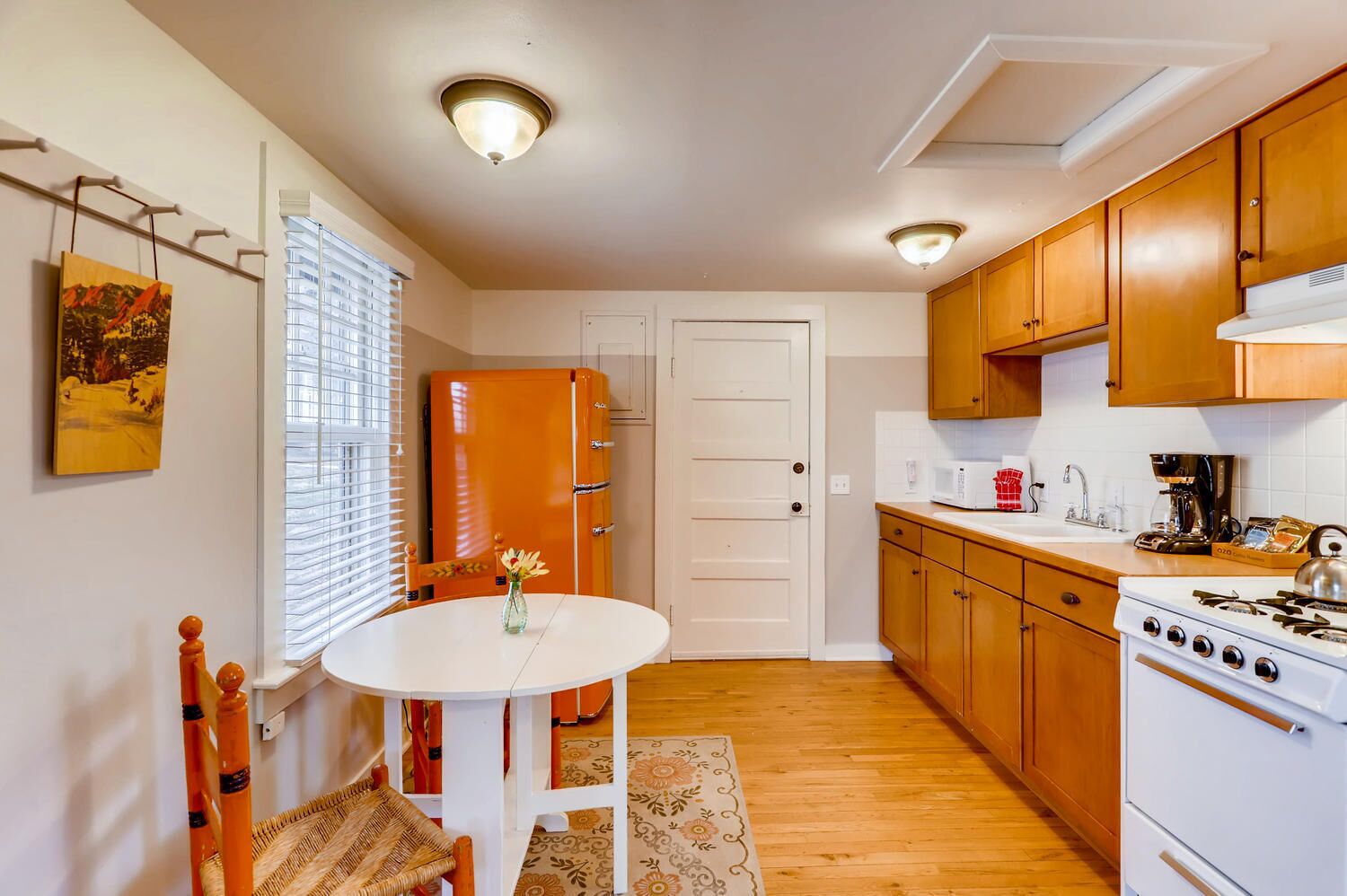 A compact kitchen with a white stove, wooden cabinets, a dining table with chairs, and a window with blinds.