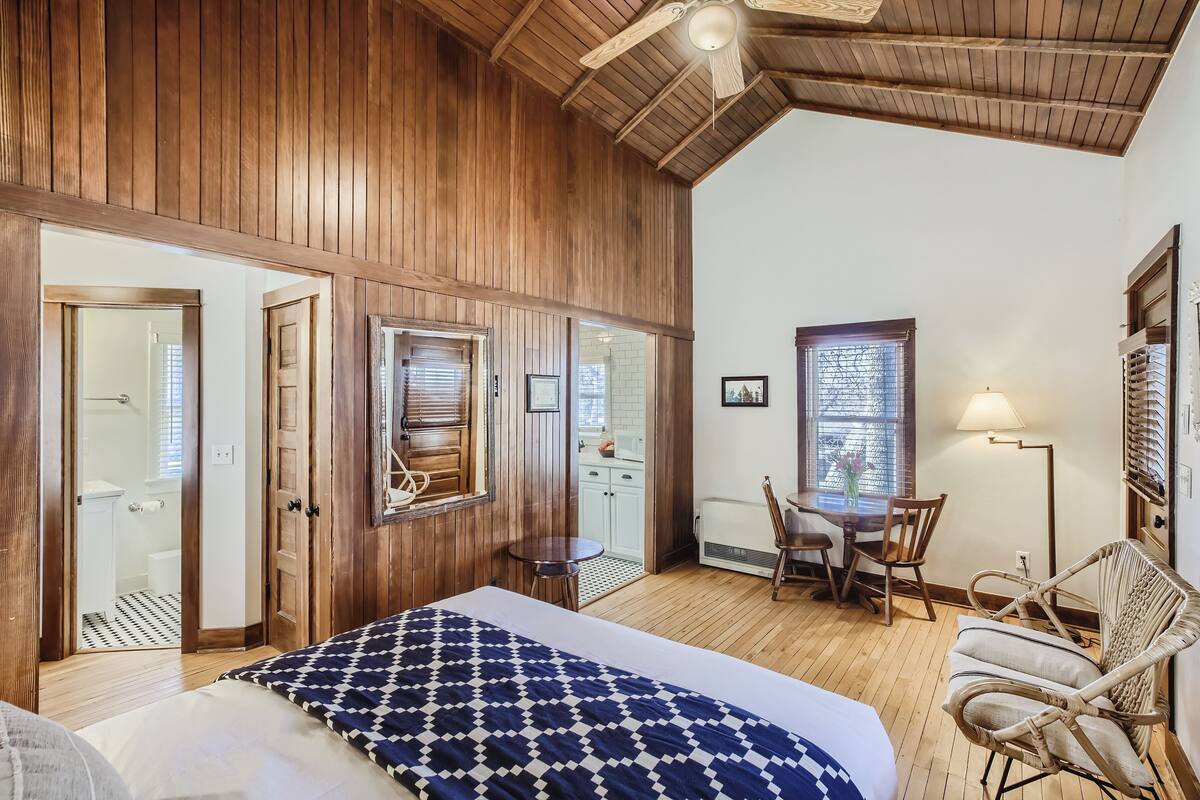 A room with a wooden ceiling, a bed with a patterned blanket, a dining table with chairs, and a lamp.