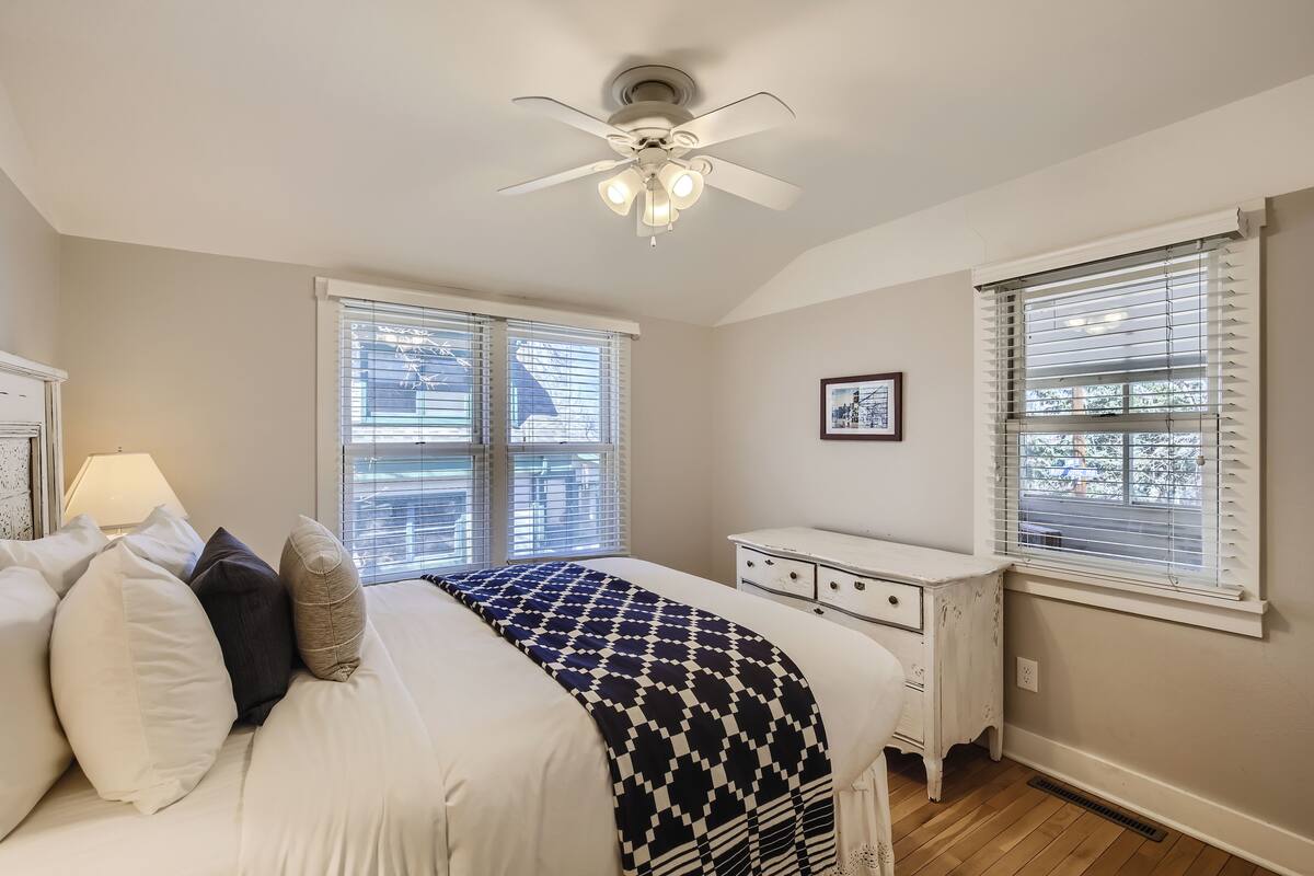 A bedroom with a bed, a ceiling fan, two windows with blinds, a bedside table, and a nightstand.
