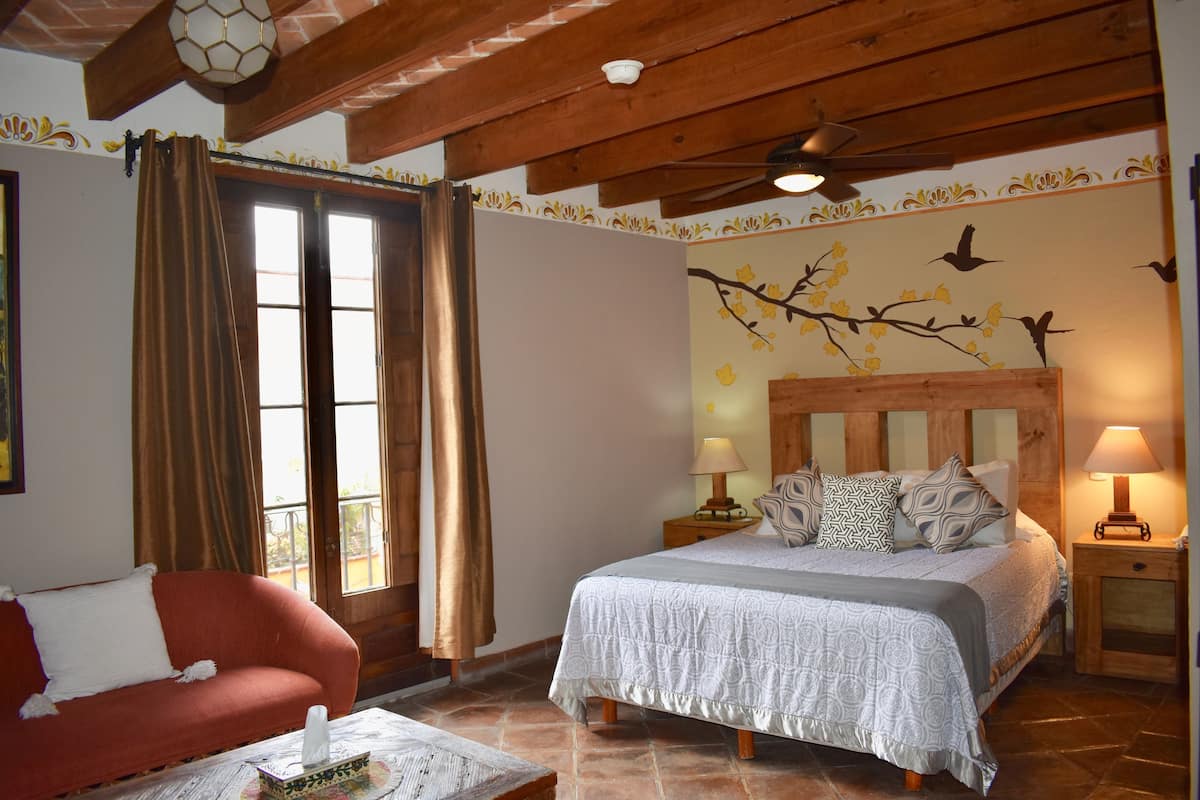 A bedroom with a bed, a red sofa, a wooden ceiling, and a wall with a branch and bird mural.