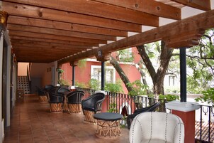 Un área de estar al aire libre techada con sillas y mesas de mimbre, un edificio rojo al fondo y un árbol.