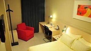 Een hotelkamer met een bed, een bureau met een stoel, een rode fauteuil, een lamp en een schilderij aan de muur.