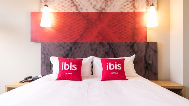 A hotel room with a large bed, two red pillows with "ibis Hotels" branding, a marble headboard, and two pendant lights. - Ibis Harbin Hongqi Street Hotel (Harbin)