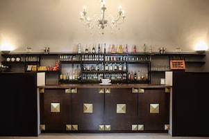 Angolo bar con un bancone in legno scuro, scaffali pieni di bottiglie e bicchieri, e un lampadario.