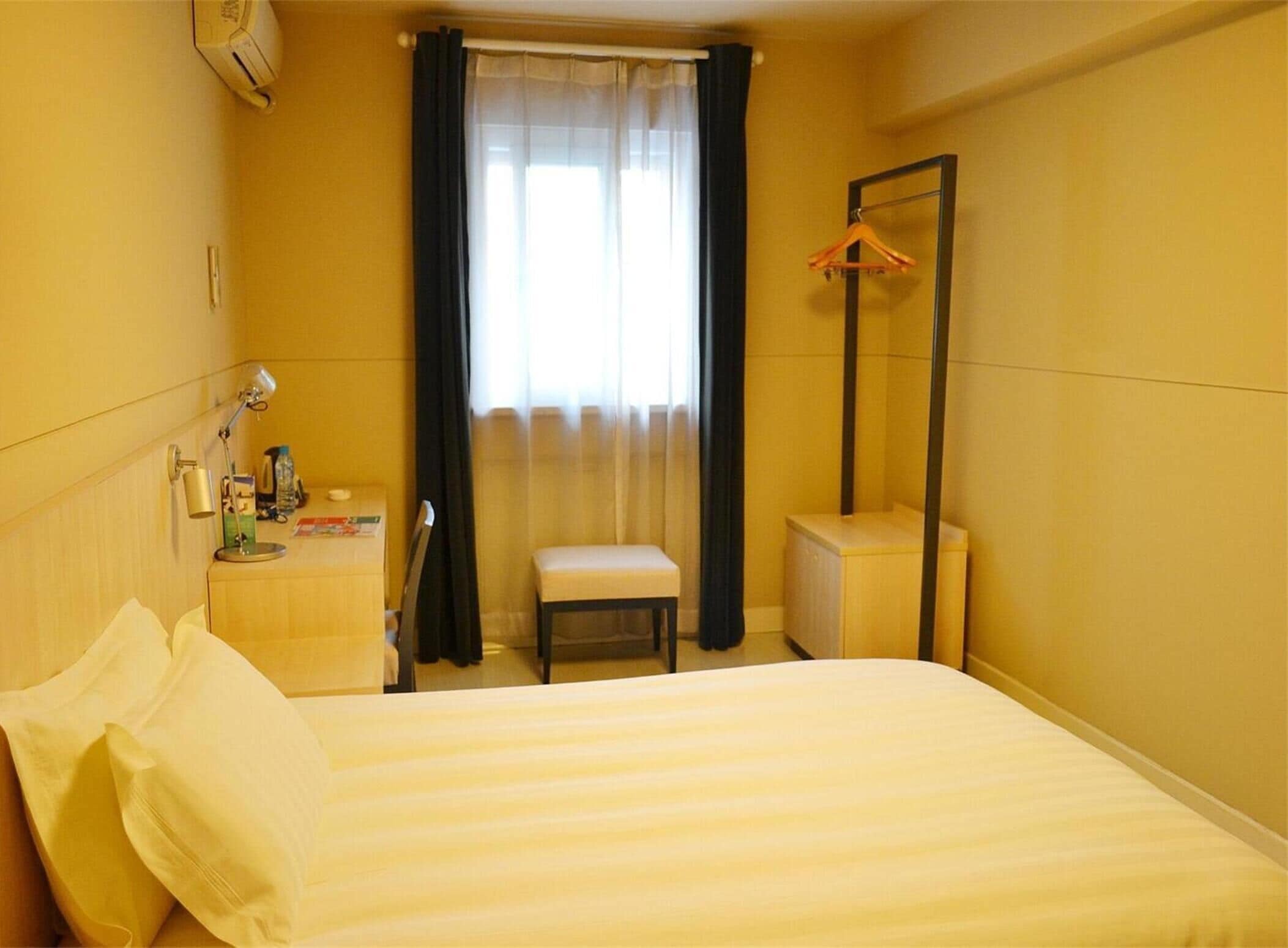 Ett hotellrum med en säng, en stol, ett litet bord med en lampa, en spegel och ett fönster med gardiner.
