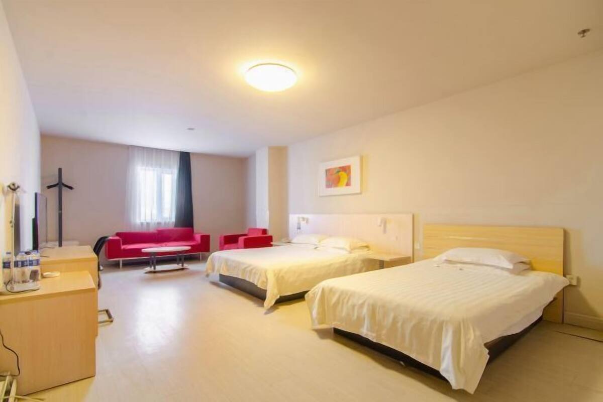 A hotel room with two beds, a desk, a red sofa, and a window with curtains.