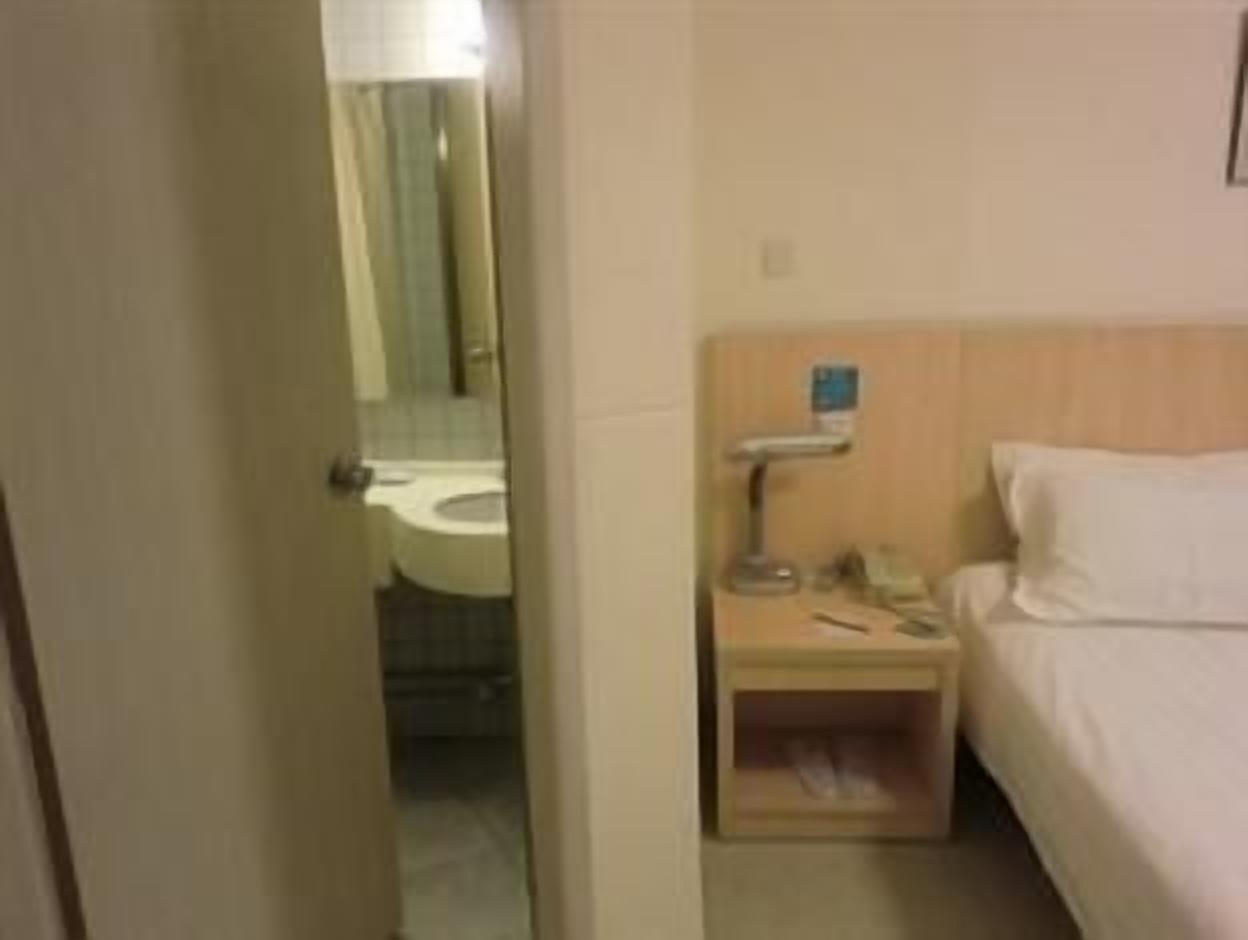 Habitación de hotel con cama, mesita de noche con lámpara y baño visible a través de una puerta abierta.
