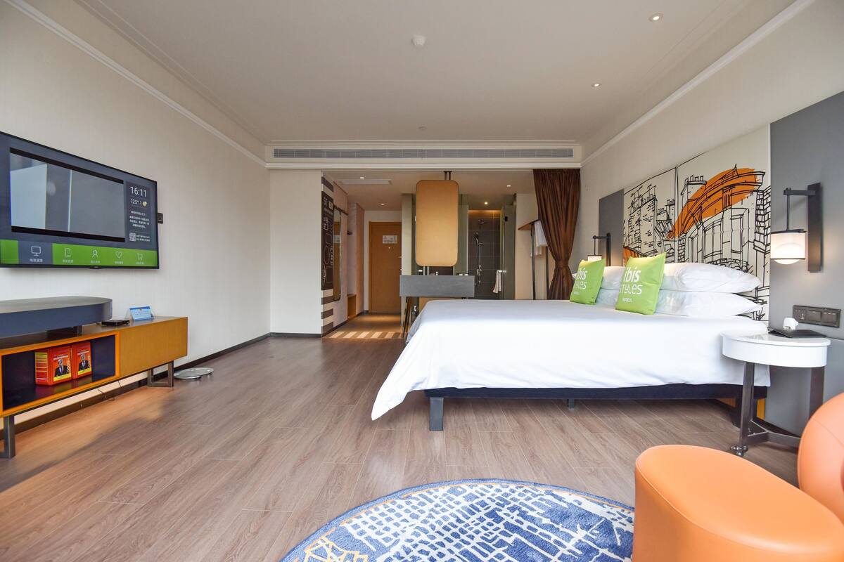 Ein Hotelzimmer mit Bett, einem an der Wand befestigten Fernseher, einem Schreibtisch mit Laptop, einem kleinen runden Teppich und einem Sessel.
