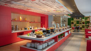 A modern cafe with a red and white color scheme, featuring a long counter with various food and drink items, a bookshelf with books and plants, and a ceiling with decorative patterns.