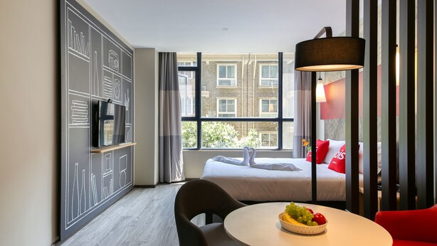 A hotel room with a bed, a TV, a dining table with fruit, and a red chair.