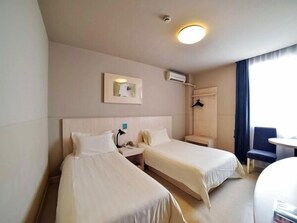 Une chambre d’hôtel avec deux lits simples, une table de chevet, une chaise, une petite table, un tableau au mur et une fenêtre avec des rideaux.