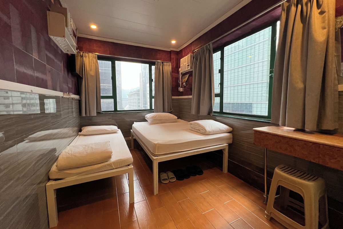 A hotel room with two single beds, a small stool, a window with curtains, and a view of buildings outside.