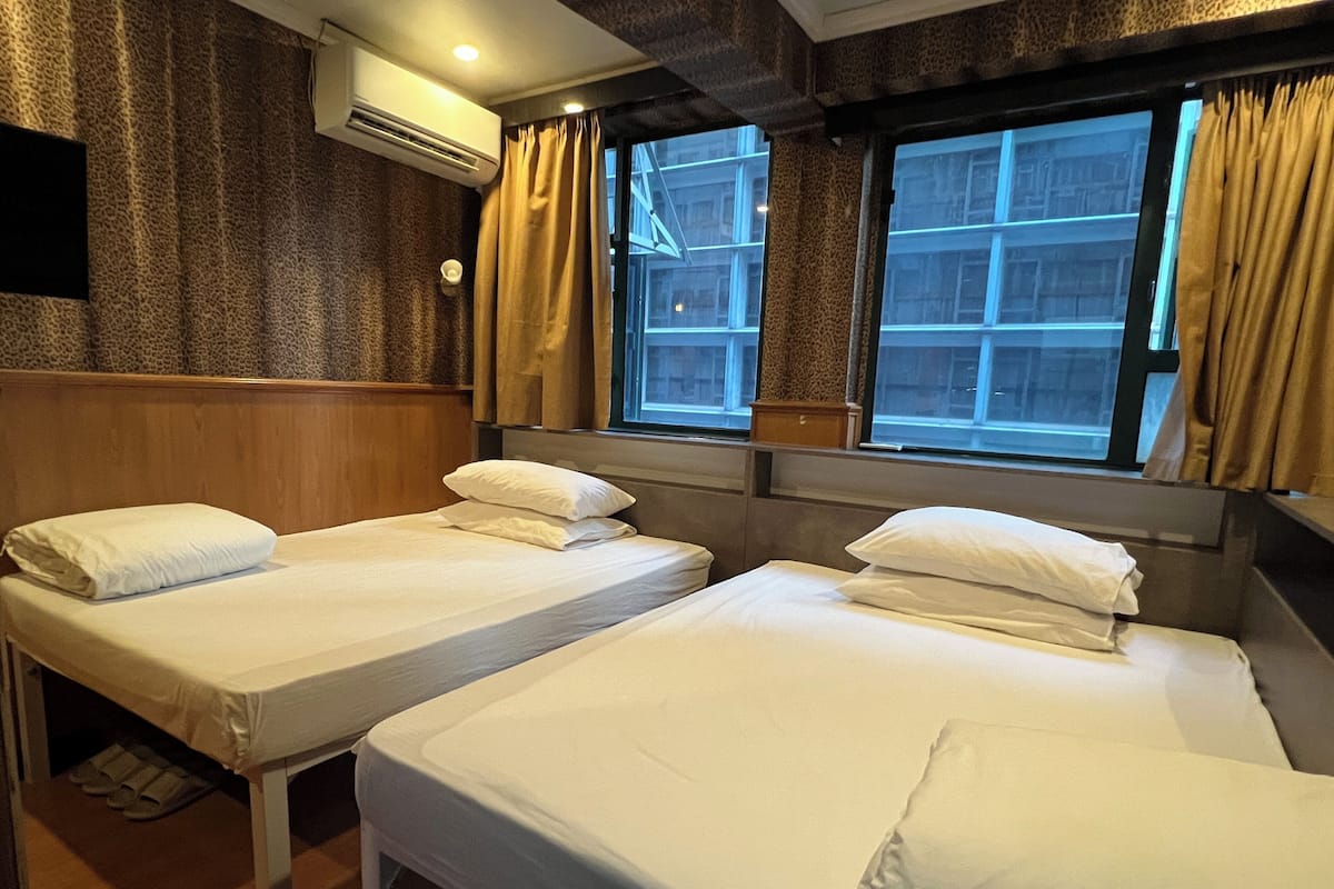 A hotel room with two single beds, a wooden headboard, an air conditioning unit, and a window with curtains.
