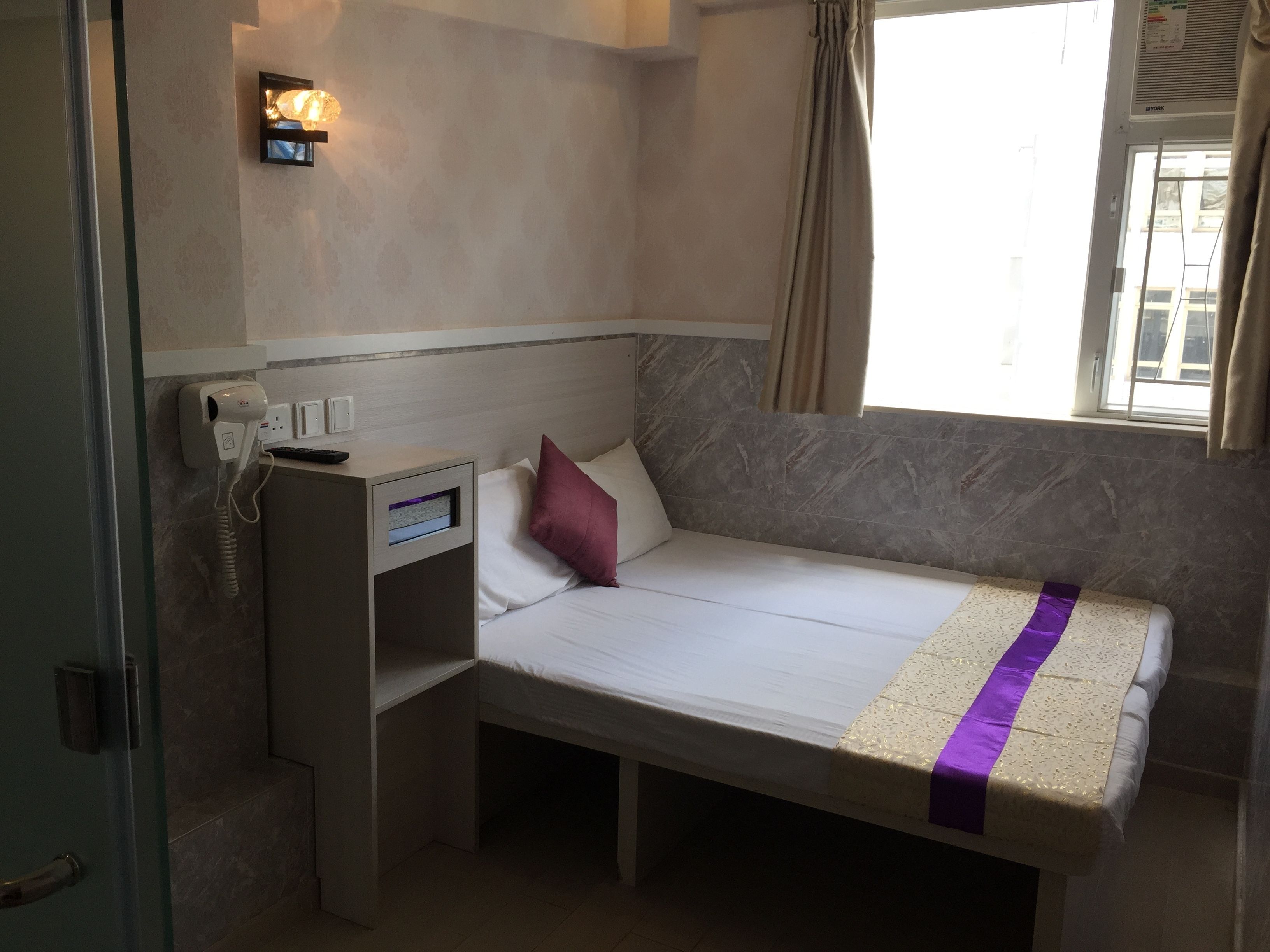 A compact hotel room with a single bed, a bedside table, a hairdryer, and a window with curtains.