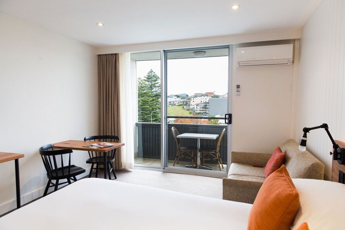 Une chambre d’hôtel comprenant un lit, un bureau avec une chaise, un balcon avec une table et des chaises, et une vue sur l’extérieur.