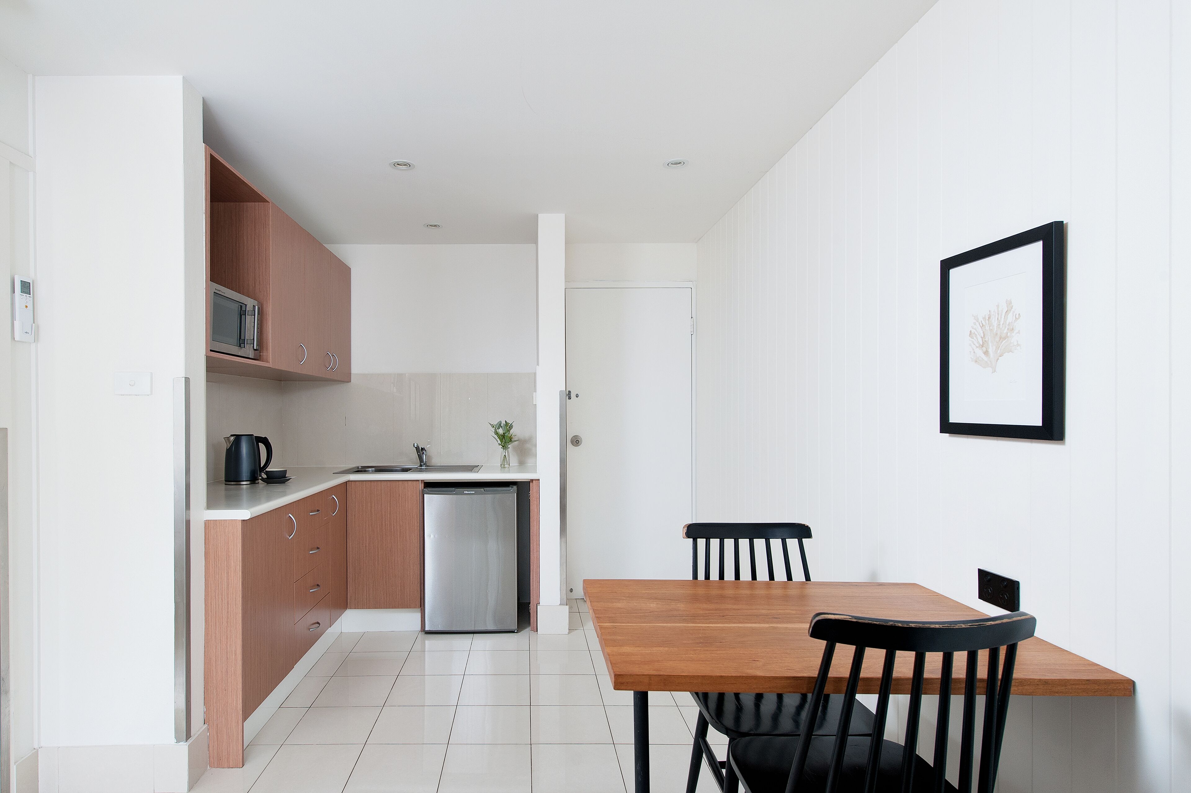 A modern kitchen with a microwave, kettle, and dishwasher, a wooden dining table with chairs, and a framed picture on the wall.