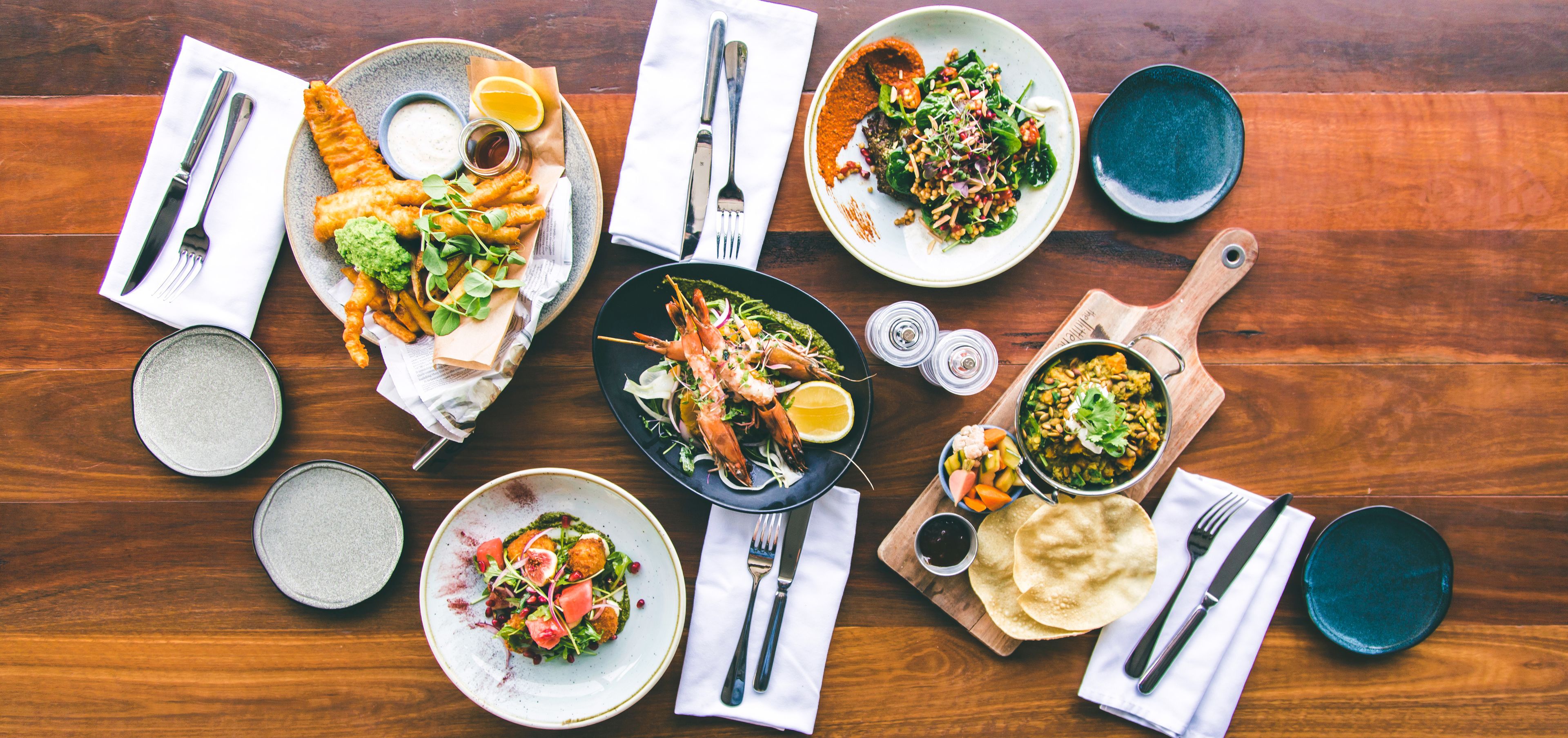 A variety of dishes on a wooden table, including seafood, salads, and flatbreads, with cutlery and condiments arranged around them.