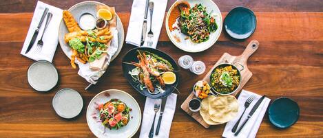 A variety of dishes on a wooden table, including seafood, salads, and flatbreads, with cutlery and condiments arranged around them.