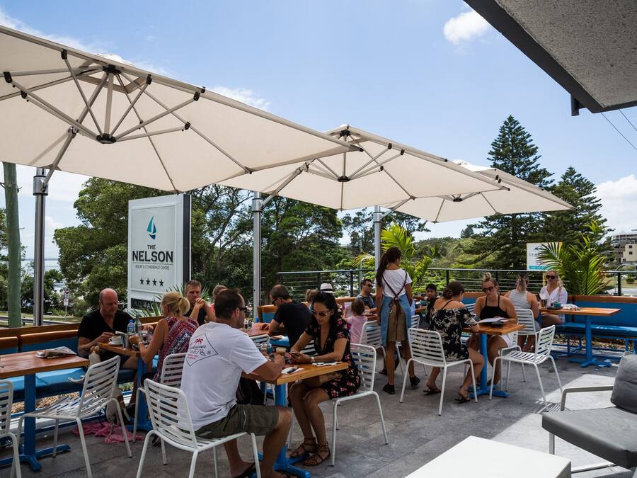 An outdoor seating area with tables and chairs under umbrellas, a sign for The Nelson Resort & Conference Centre, and a view of trees and buildings in the background.