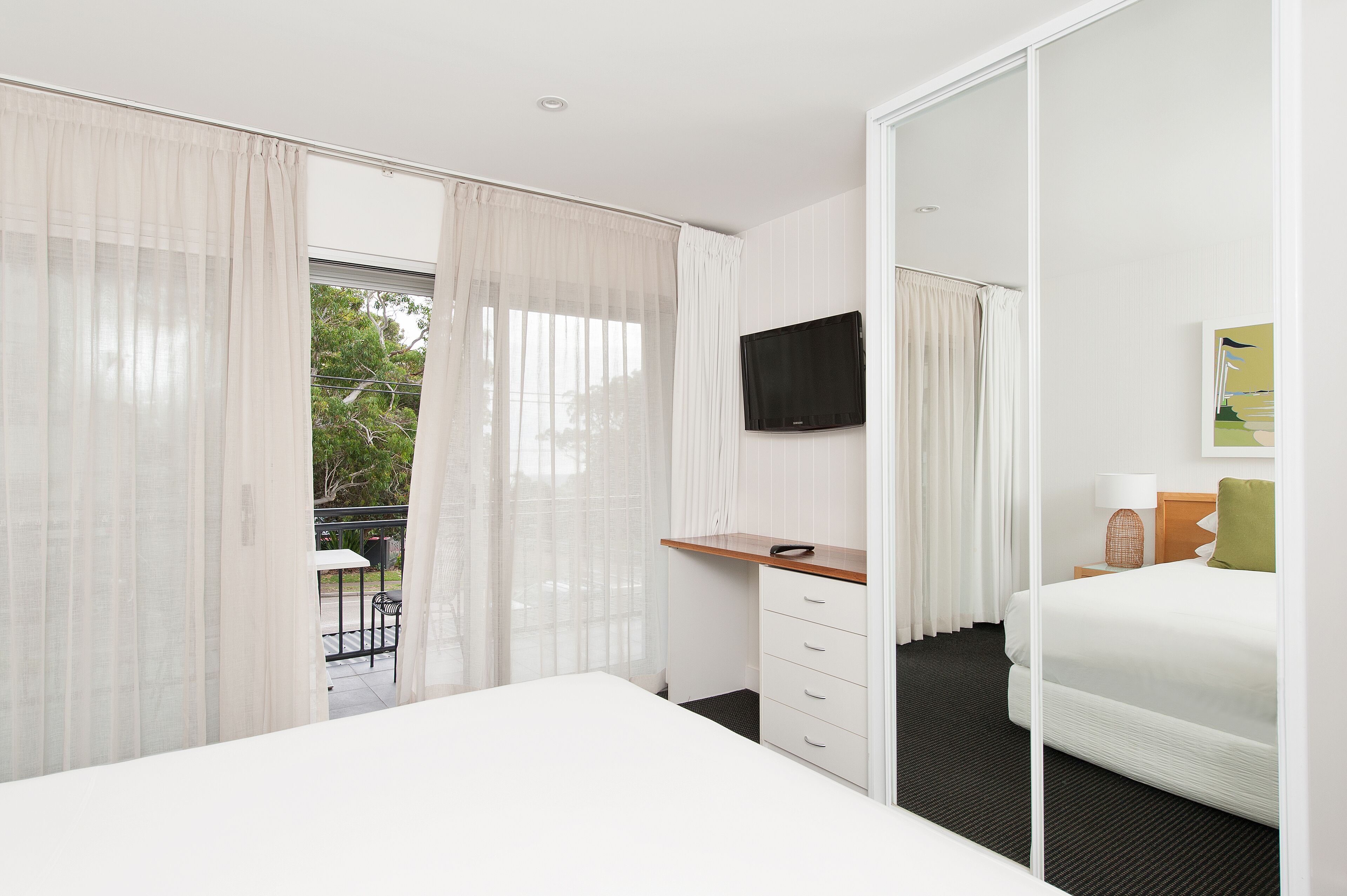 A hotel room with a bed, a desk, a chair, a TV, and a balcony with a view of trees.